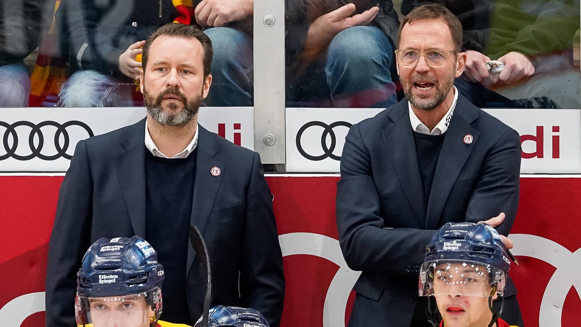 Roger Hansson (r) und sein Co-Trainer Thomas Dolak (l) stehen nebeneinander im Eisstadion. Vor ihnen sitzen zwei Eishockey-Spieler. Roger Hansson (r) und sein Co-Trainer Thomas Dolak (l) stehen nebeneinander im Eisstadion. Vor ihnen sitzen zwei Eishockey-Spieler.