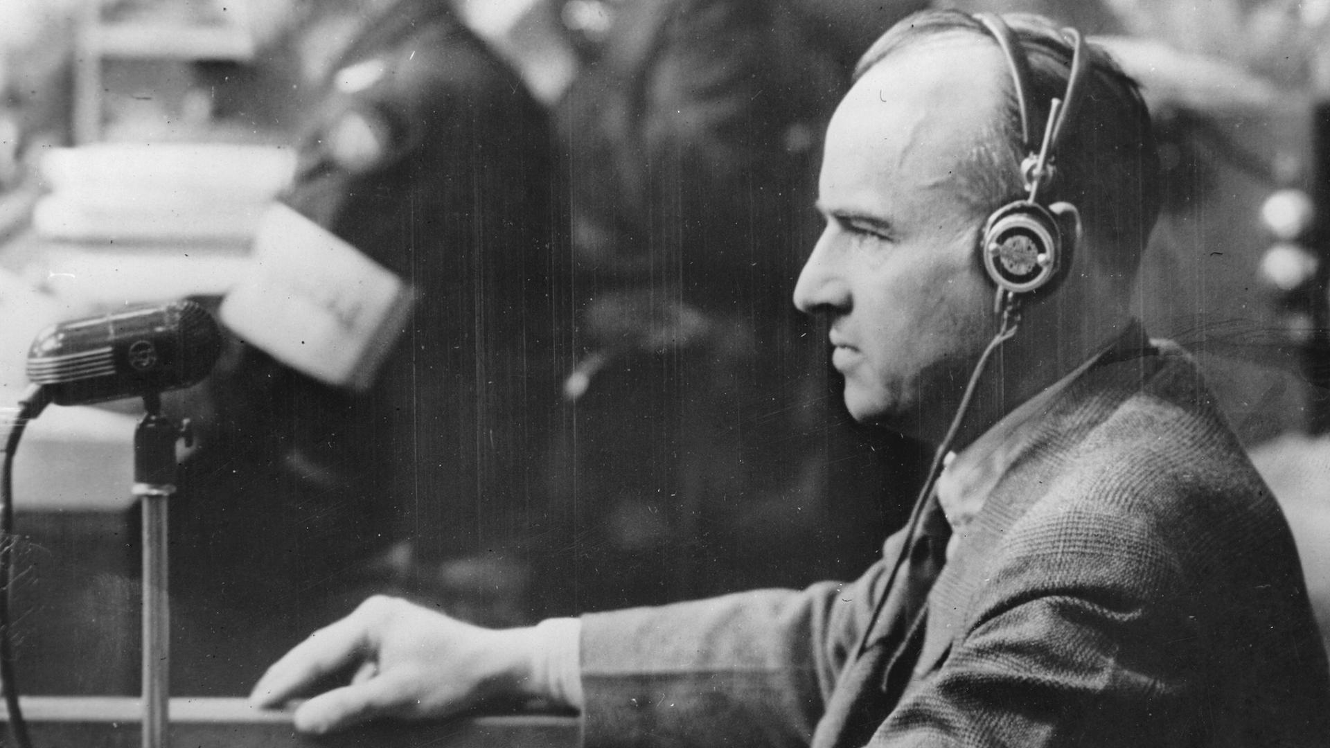 Nationalsozialist Hans Frank beim Nürnberger Prozess. Er trägt einen Kopfhörer und steht vor einem Mikrofon.