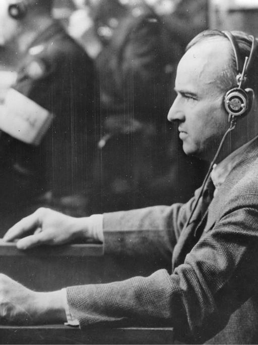 Nationalsozialist Hans Frank beim Nürnberger Prozess. Er trägt einen Kopfhörer und steht vor einem Mikrofon.