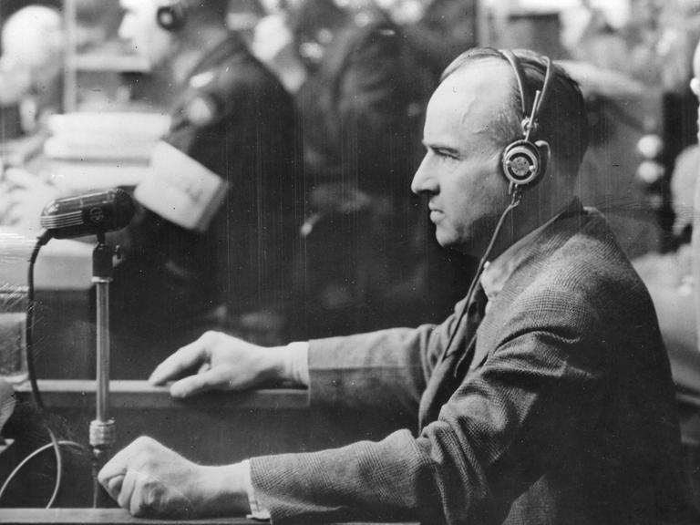 Nationalsozialist Hans Frank beim Nürnberger Prozess. Er trägt einen Kopfhörer und steht vor einem Mikrofon.