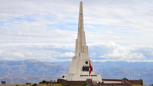 Ein weißer Obelisk steht auf einer Anhöhe. Im Hintergrund sind Bergketten zu sehen. 