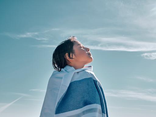 Ein Junge, eingewickelt in eine Decke, steht mit geschlossenen Augen vor blauem Himmel und atmet bewusst ein und aus. Ein Junge, eingewickelt in eine Decke, steht mit geschlossenen Augen vor blauem Himmel und atmet bewusst ein und aus.