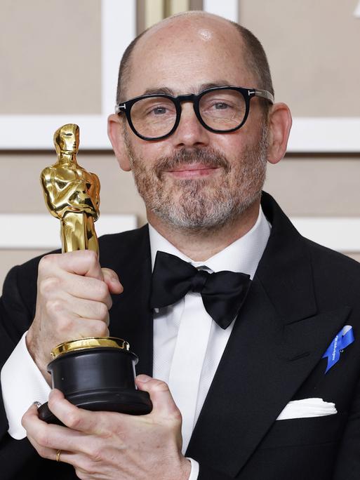 Edward Berger zeigt die goldene Oscar-Figur in die Kamera. Er lächelt verhalten. Der Hintergrund ist hell.