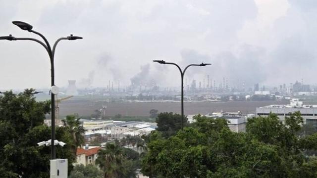 Rauch steigt über den entfernten Türmen der Ölraffinerie von Haifa auf. Im Vordergrund sind Bäume und Häuser.