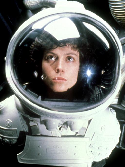 Sigourney Weaver als Officer Ellen Ripley trägt einen Raumanzug und schaut durch das Sichtfenster ihres Helms nach oben.