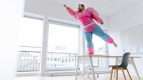 Ein Mann in einem aufblasbaren Superheldenkostüm mit großen Muskeln steht auf einem Stuhl und nimmt eine Pose ein, als könne er fliegen.