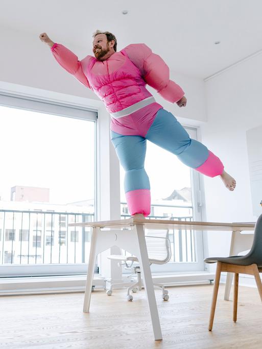 Ein Mann in einem aufblasbaren Superheldenkostüm mit großen Muskeln steht auf einem Stuhl und nimmt eine Pose ein, als könne er fliegen.