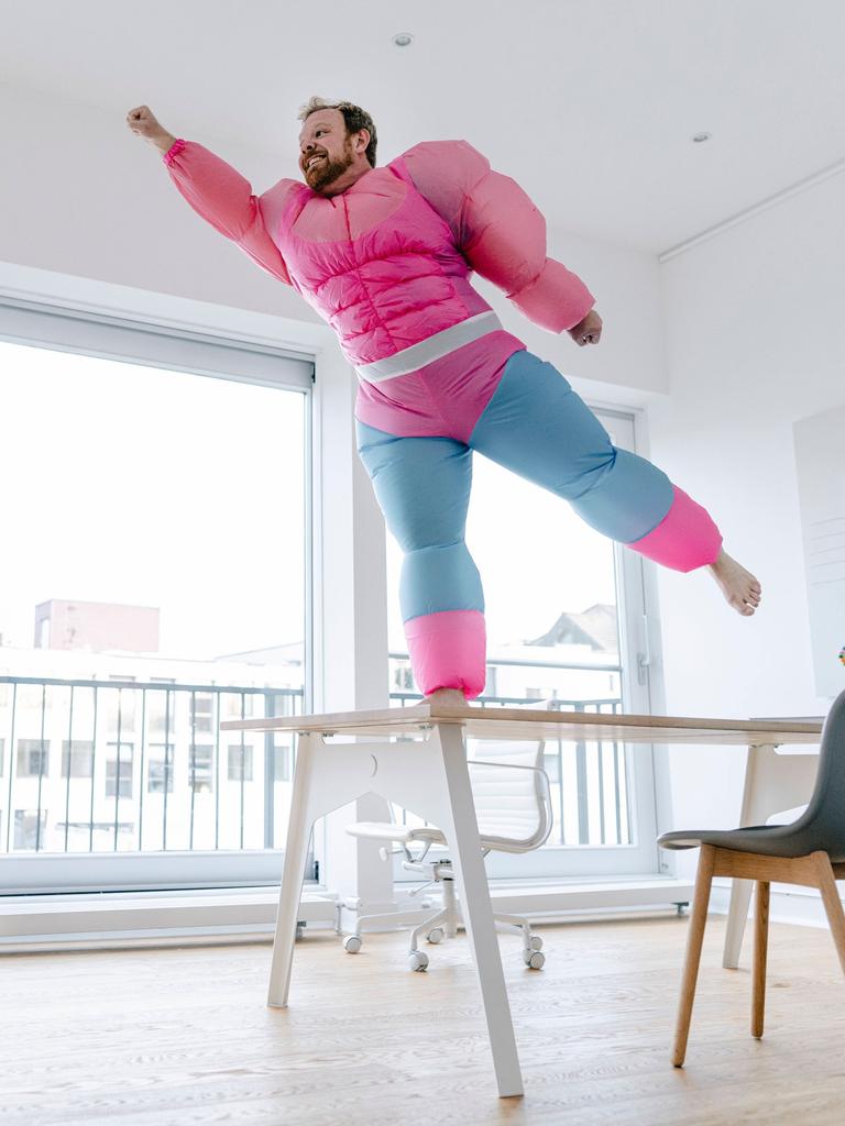 Ein Mann in einem aufblasbaren Superheldenkostüm mit großen Muskeln steht auf einem Stuhl und nimmt eine Pose ein, als könne er fliegen.