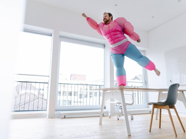 Ein Mann in einem aufblasbaren Superheldenkostüm mit großen Muskeln steht auf einem Stuhl und nimmt eine Pose ein, als könne er fliegen.
