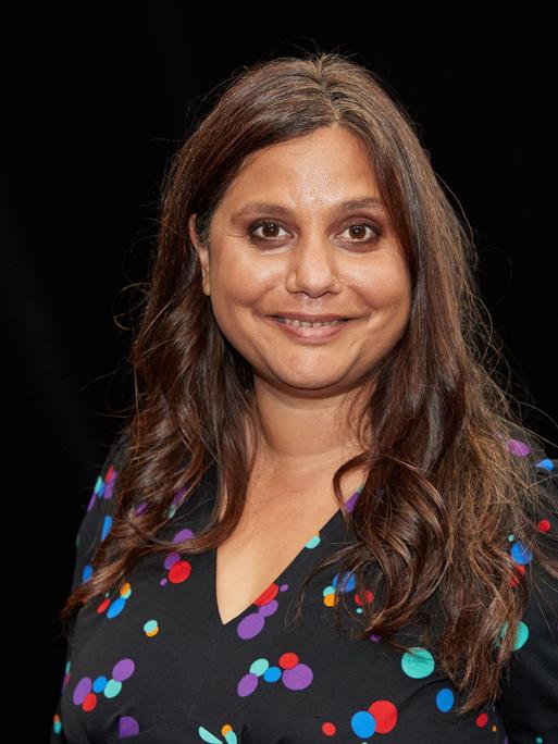 Mithu Sanyal, Kulturwissenschaftlerin, blickt in die Kamera. Sie trägt ein dunkles, bunt gepunktetes Oberteil, sie hat lange, schwarze Haare.