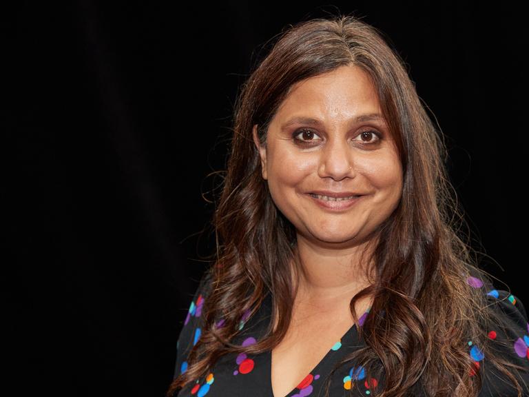 Mithu Sanyal, Kulturwissenschaftlerin, blickt in die Kamera. Sie trägt ein dunkles, bunt gepunktetes Oberteil, sie hat lange, schwarze Haare.