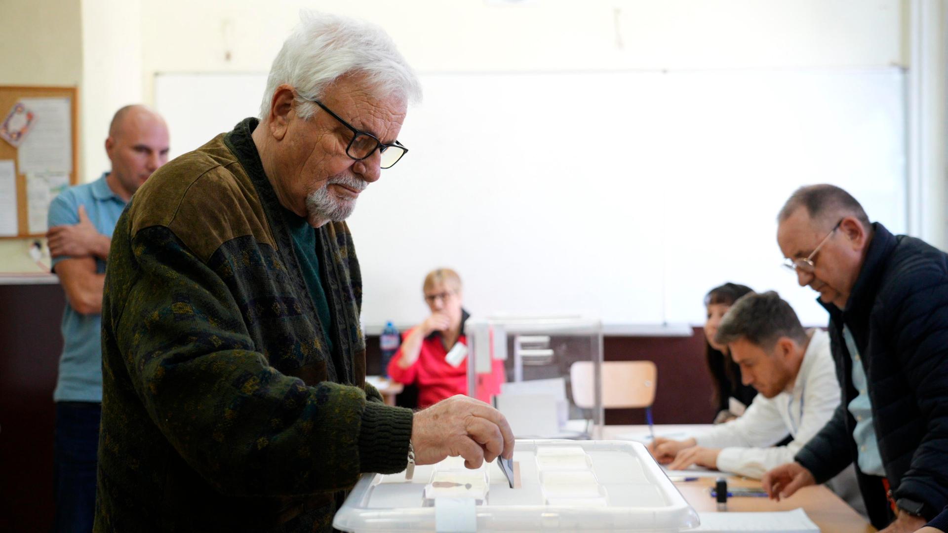 Ein Mann mit grauem Bart und Brille wirft einen Stimmzettel in eine Wahlurne. Das Wahllokal ist in der bulgarischen Hauptstadt Sofia. 
