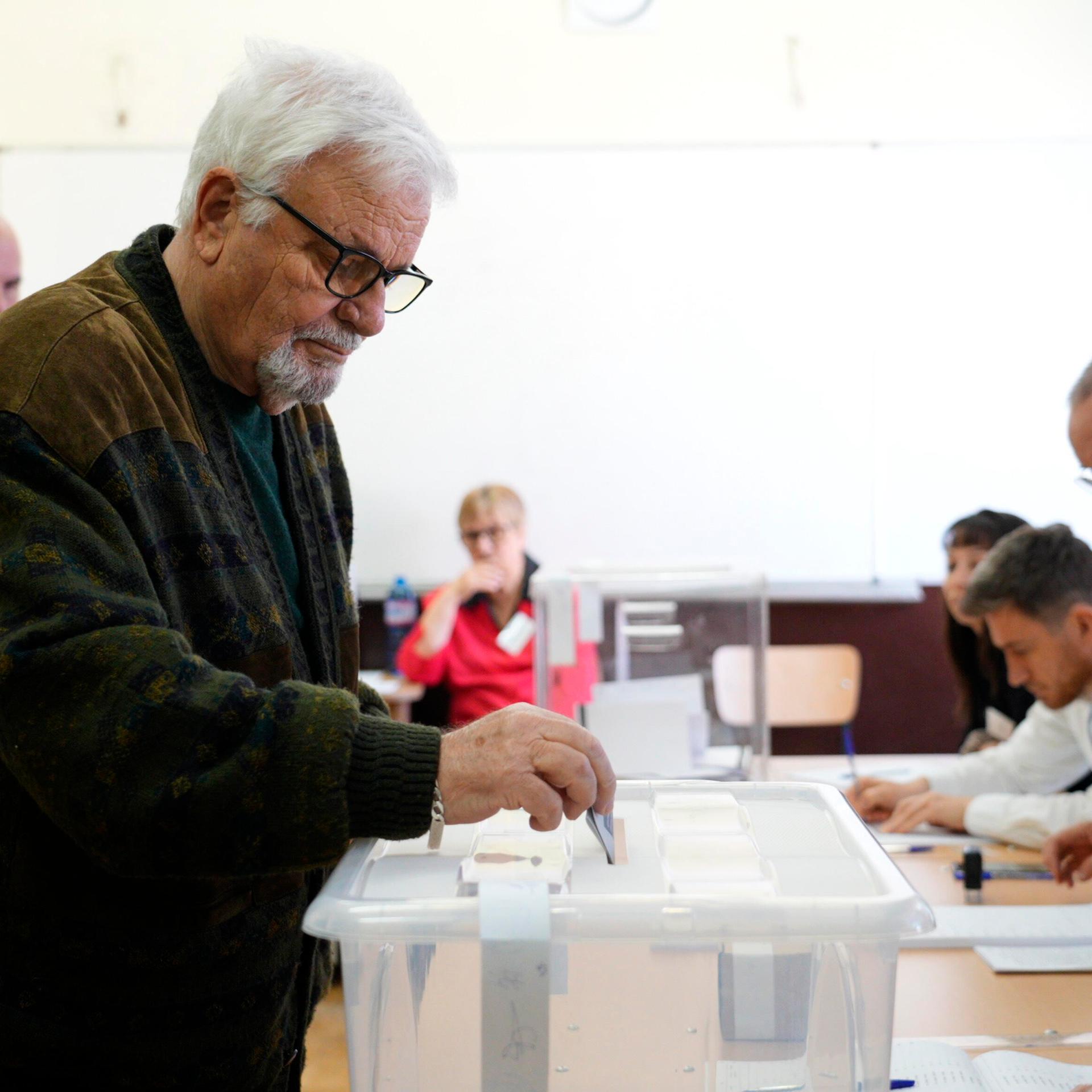 Ein Mann mit grauem Bart und Brille wirft einen Stimmzettel in eine Wahlurne. Das Wahllokal ist in der bulgarischen Hauptstadt Sofia. 