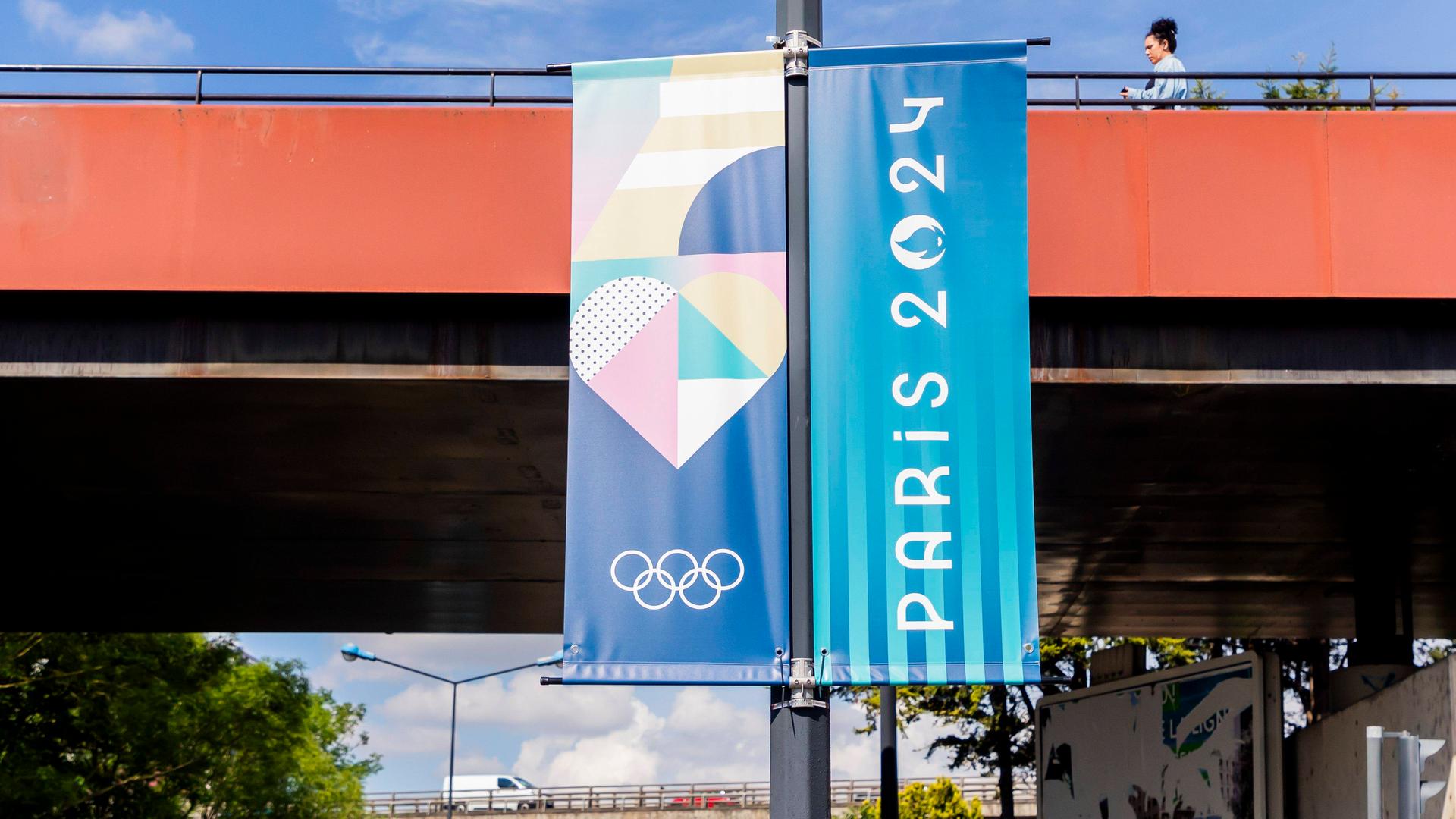 Zu sehen ist eine Fahne, im Stil zweigeteilt: Die eine Seite zeigt den Schriftzug "Paris 2024", die anderen Seite zeigt die Olympischen Ringe und bunte Formen. Die Fahne hängt an einer Straßenlaterne an einer viel befahrenen Straße.
