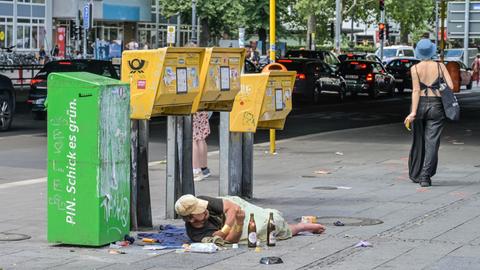 Obdachloser Alkoholiker liegt am Bahnhof Zoo in Berlin vor Briefkästen auf dem Bürgersteig. Eine junge Frau schlendert vorbei.  