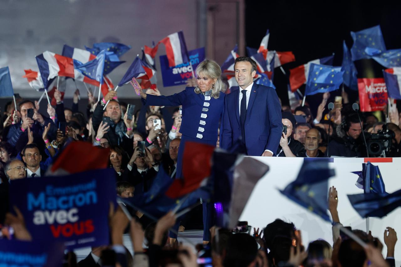 Der französische Präsident und Kandidat der Partei La Republique en Marche für die Wiederwahl, Emmanuel Macrons, und seine Frau Brigitte Macron feiern nach seinem Sieg bei den französischen Präsidentschaftswahlen auf dem Champ de Mars in Paris. Sie stehen auf einer Bühne, umgeben von Anhängern und winken lachend in die Menge.