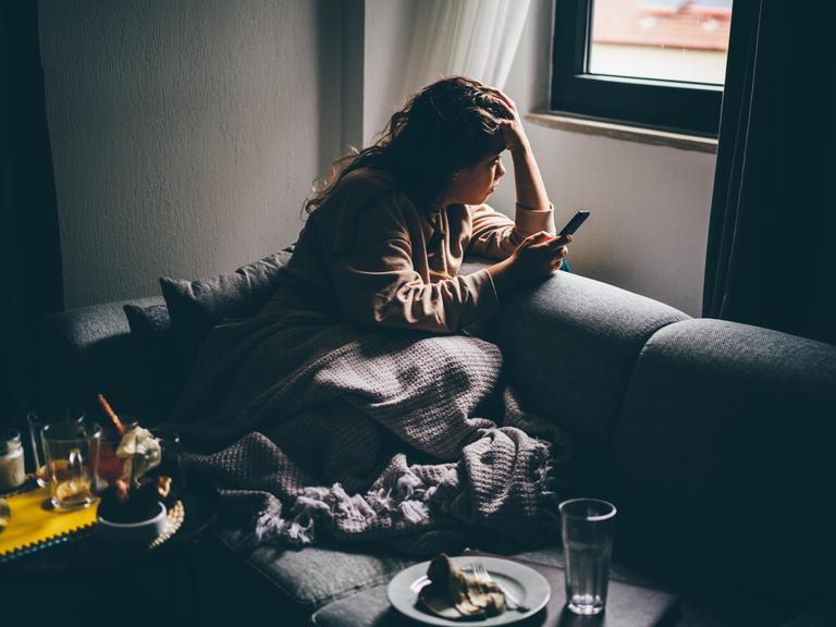 Eine junge Frau mit traurigem Gesichtsausdruck sitzt im Halbdunkel mit ihrem Smartphone in der Hand auf dem Sofa.