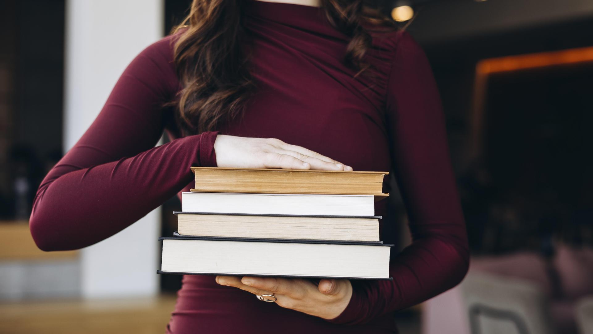 Eine junge Frau in einem kastanienbraunen Oberteil hält einen Stapel Bücher in den Händen. Ihr Kopf ist nicht zu sehen. 