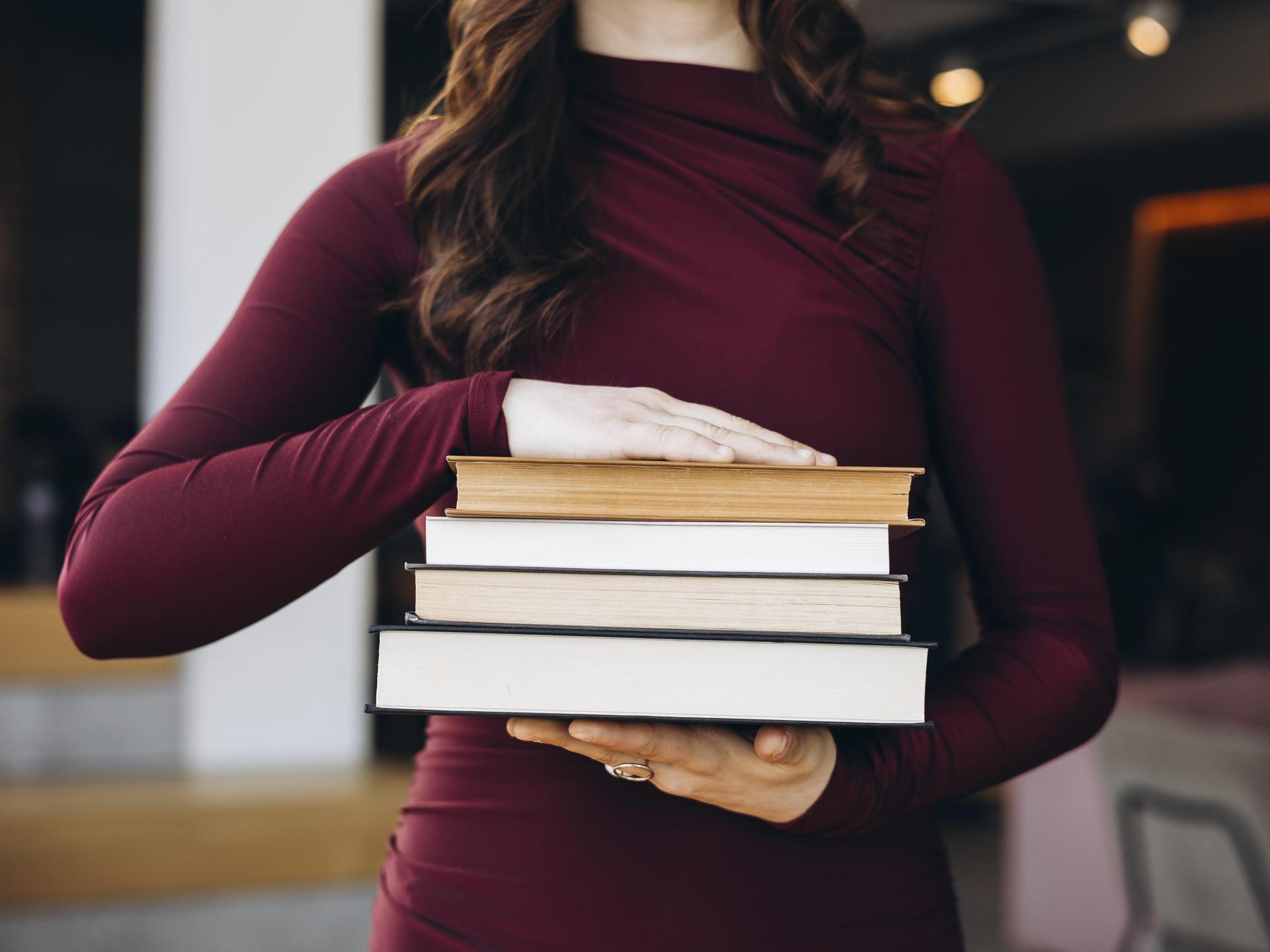 Eine junge Frau in einem kastanienbraunen Oberteil hält einen Stapel Bücher in den Händen. Ihr Kopf ist nicht zu sehen. Eine junge Frau in einem kastanienbraunen Oberteil hält einen Stapel Bücher in den Händen. Ihr Kopf ist nicht zu sehen.