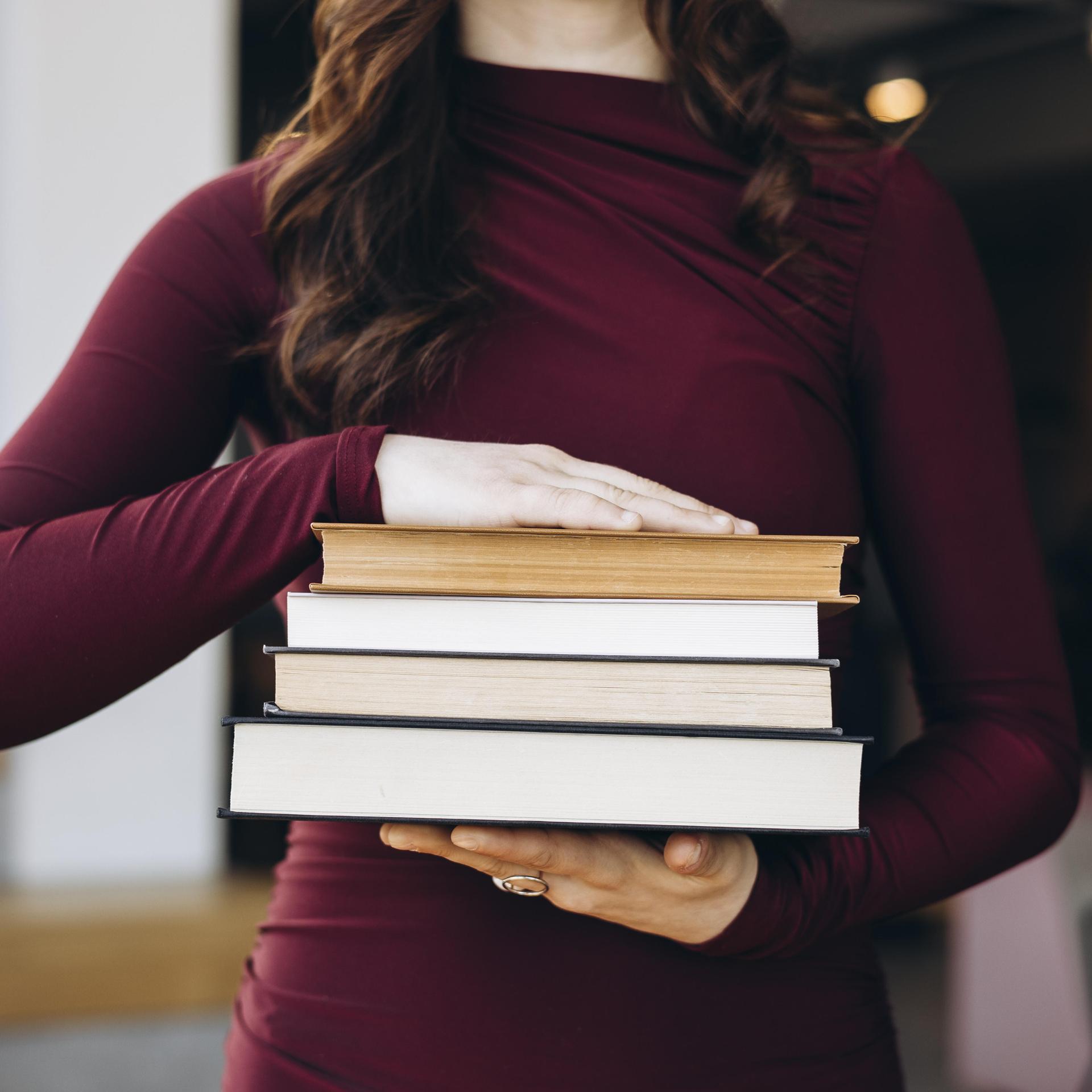 Eine junge Frau in einem kastanienbraunen Oberteil hält einen Stapel Bücher in den Händen. Ihr Kopf ist nicht zu sehen. 