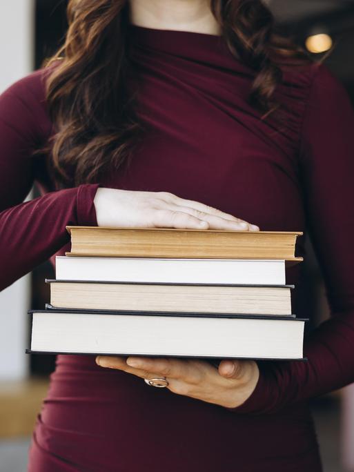 Eine junge Frau in einem kastanienbraunen Oberteil hält einen Stapel Bücher in den Händen. Ihr Kopf ist nicht zu sehen. 