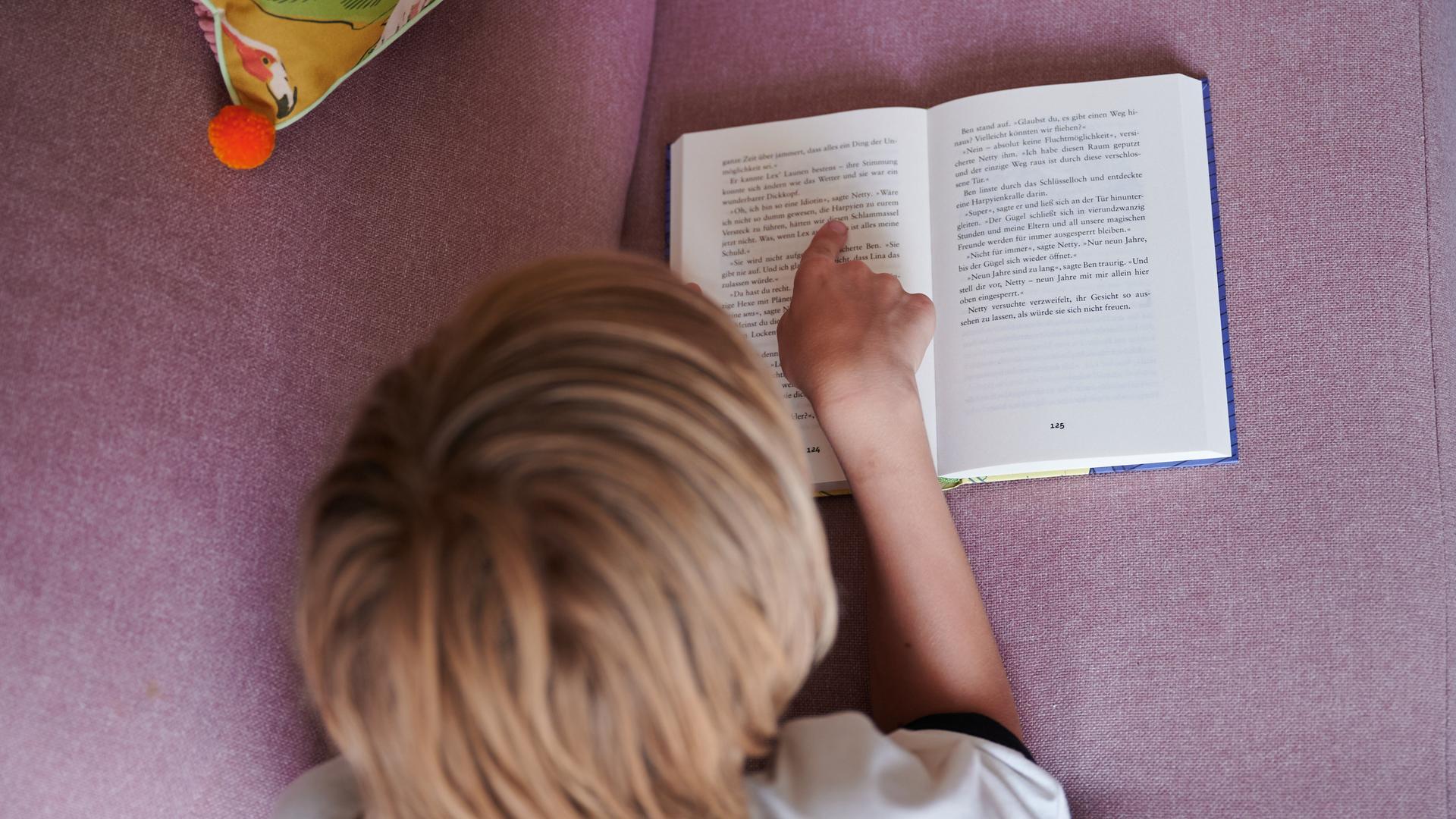 Ein Kind liest mit dem Finger auf einer Zeile in einem aufgeschlagenen Buch, während es auf dem Bauch auf einer Couch liegt. (gestellte Szene) Ein Kind liest mit dem Finger auf einer Zeile in einem aufgeschlagenen Buch, während es auf dem Bauch auf einer Couch liegt. (gestellte Szene)