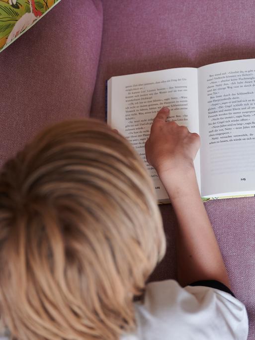 Ein Kind liest mit dem Finger auf einer Zeile in einem aufgeschlagenen Buch, während es auf dem Bauch auf einer Couch liegt. (gestellte Szene)