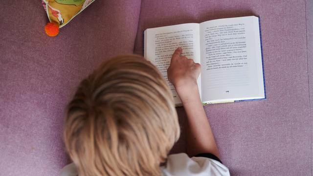 Ein Kind liest mit dem Finger auf einer Zeile in einem aufgeschlagenen Buch, während es auf dem Bauch auf einer Couch liegt. (gestellte Szene)