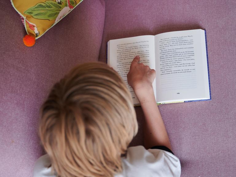 Ein Kind liest mit dem Finger auf einer Zeile in einem aufgeschlagenen Buch, während es auf dem Bauch auf einer Couch liegt. (gestellte Szene)