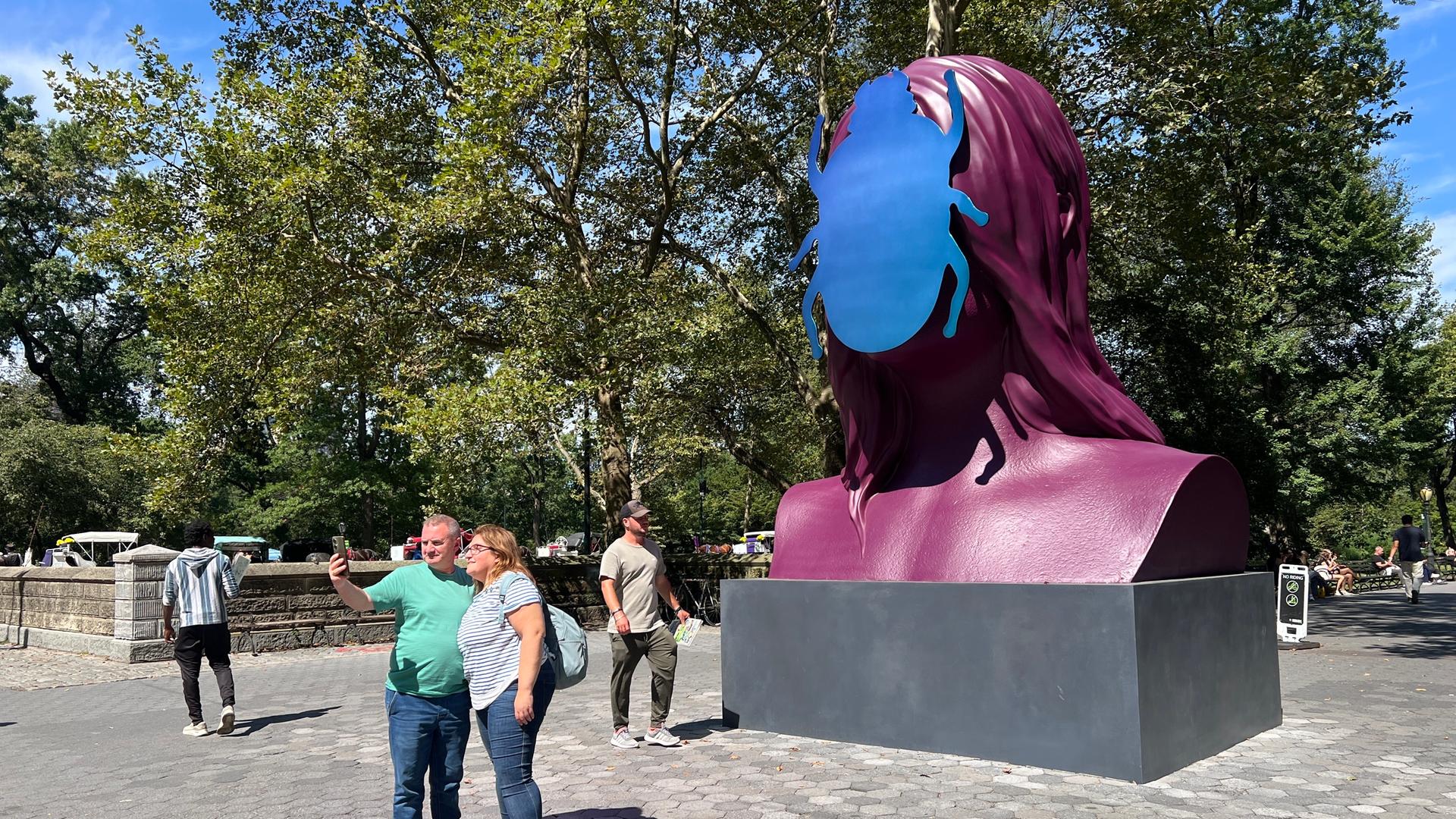 Besucher machen ein Selfie vor einer großen Skulptur einer Frauenbüste mit einem Käfer anstelle des Kopfes