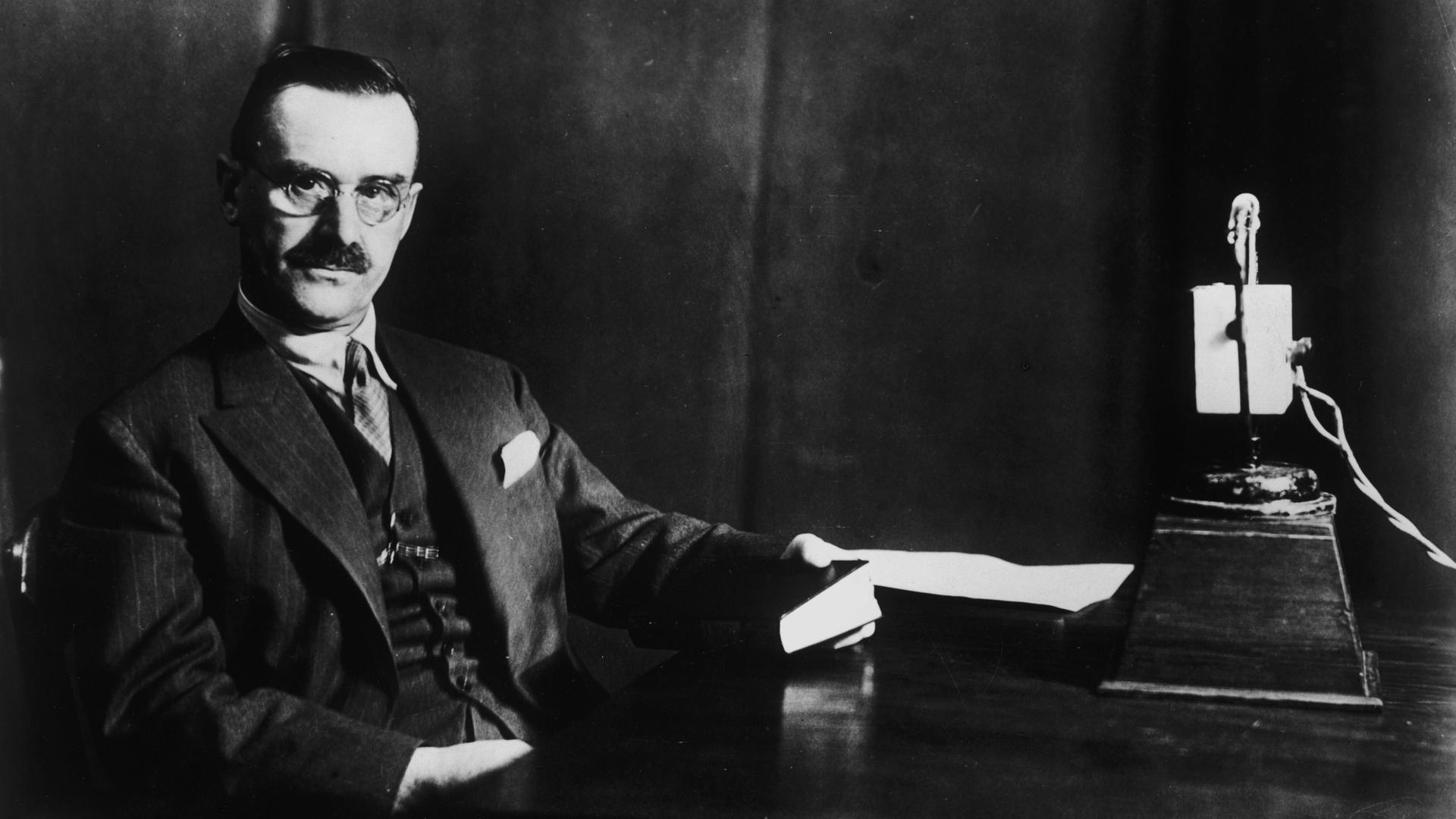 Der Schriftsteller Thomas Mann sitzt an einem Schreibtisch, in seiner Hand ein Buch. Rechts auf dem Schreibtisch steht ein Mikrofon. Der Schriftsteller Thomas Mann sitzt an einem Schreibtisch, in seiner Hand ein Buch. Rechts auf dem Schreibtisch steht ein Mikrofon.