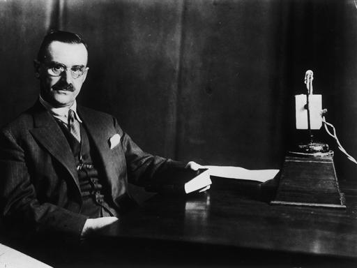 Der Schriftsteller Thomas Mann sitzt an einem Schreibtisch, in seiner Hand ein Buch. Rechts auf dem Schreibtisch steht ein Mikrofon.