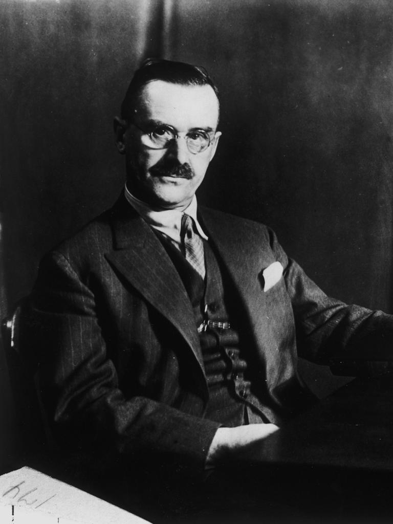 Der Schriftsteller Thomas Mann sitzt an einem Schreibtisch, in seiner Hand ein Buch. Rechts auf dem Schreibtisch steht ein Mikrofon.