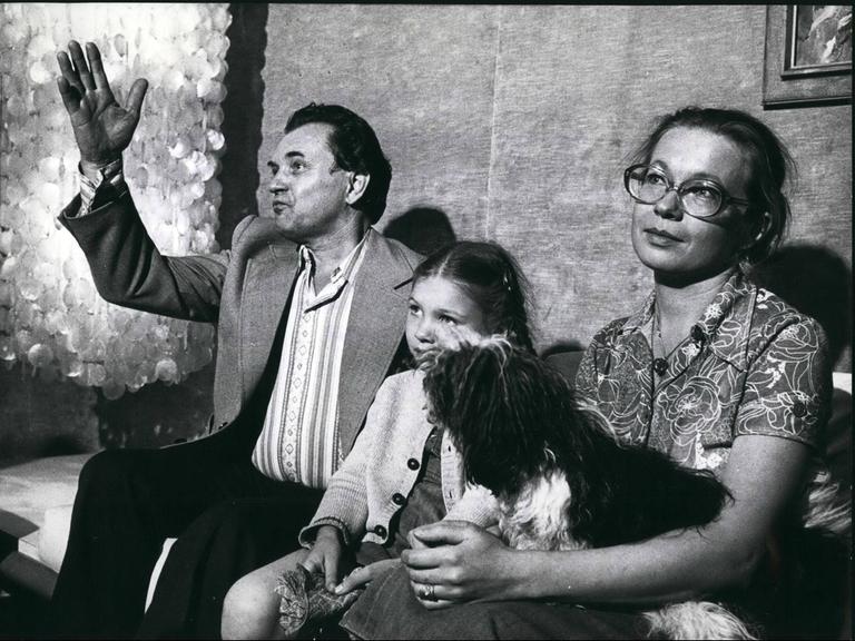 Auf dem Schwarz-weiß-Bild sind ein Mann, ein Kind und eine Frau zu sehen, die auf einer Bank sitzen. Auf dem Schwarz-weiß-Bild sind ein Mann, ein Kind und eine Frau zu sehen, die auf einer Bank sitzen.