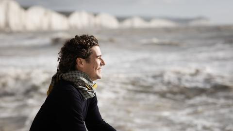 Robin Ticciati sitzt an der Hafenkante vor aufgewühltem Meer und lacht in den Wind hinein. 