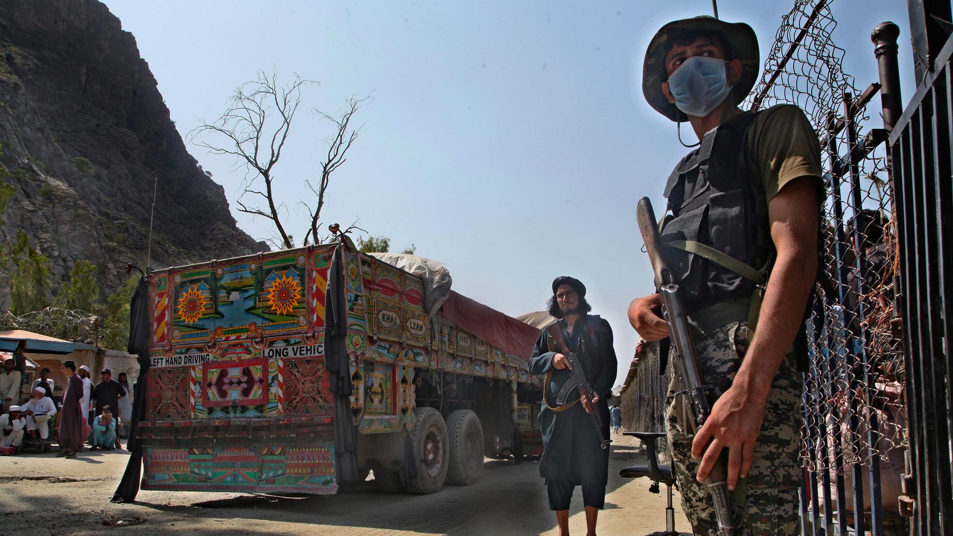 21.08.2021, Pakistan, Torkham: Ein pakistanischer Soldat (r) und Kämpfer der Taliban stehen Wache an einem Grenzübergang zwischen Pakistan und Afghanistan in der Provinz Khyber Pakhtunkhwa auf den jeweiligen Seiten der Länder.