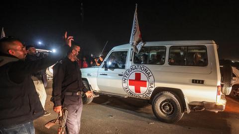 Zu sehen ist ein Jeep des Roten Kreuzes. Daneben stehen mehrere Männer, einer hält ein Gewehr in der Hand.