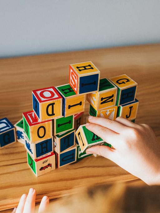 Hände stapeln Buchstabenwürfel auf einem Tisch. (Symbolbild)