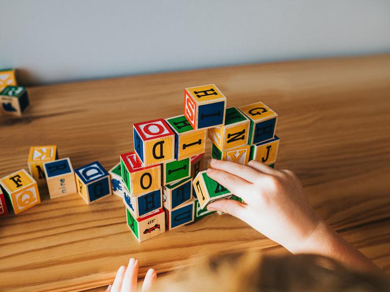 Hände stapeln Buchstabenwürfel auf einem Tisch. (Symbolbild)