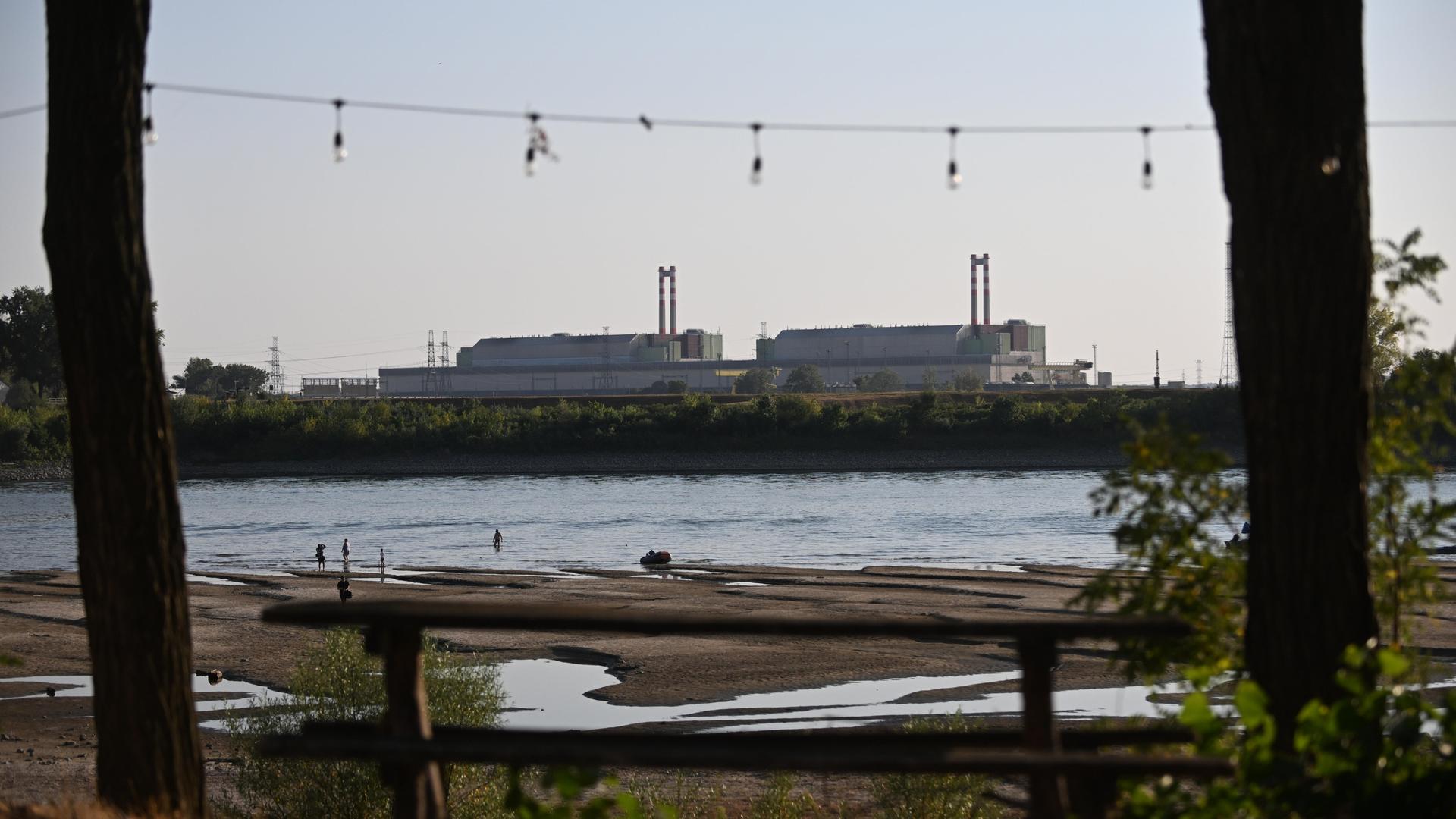 Am Ufer der Donau liegt das ungarische Atomkraftwerk Paks. Der Fluss hat einen niedrigen Pegelstand, Sandbänke treten hervor. Im Hintergrund stehen die Gebäude der Kernkraftanlage.