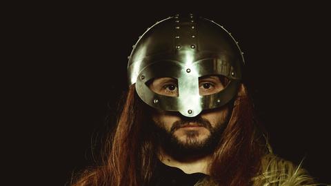 Ein Mann mit langen Haaren trägt einen eisernen Helm, der in eine eiserne Maske übergeht und blickt in die Kamera.