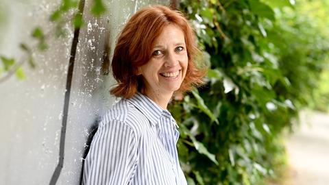 Sabine Bohlmann lacht in die Kamera. Sie hat schulterlanges rotbraunes Haar und trägt ein blauweiß gestreiftes Hemd.