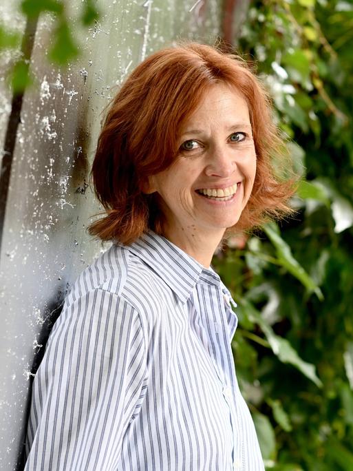 Sabine Bohlmann lacht in die Kamera. Sie hat schulterlanges rotbraunes Haar und trägt ein blauweiß gestreiftes Hemd.