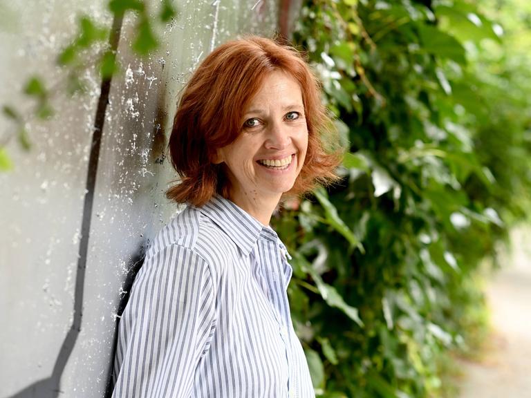 Sabine Bohlmann lacht in die Kamera. Sie hat schulterlanges rotbraunes Haar und trägt ein blauweiß gestreiftes Hemd.