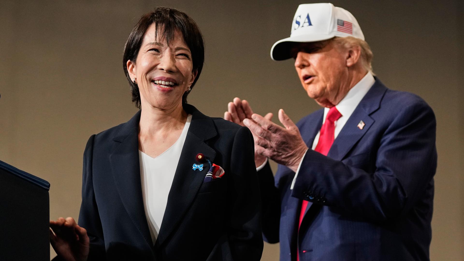 Die japanische Premierministerin Takaichi lacht, hinter ihr steht Präsident Trump und applaudiert. Die japanische Premierministerin Takaichi lacht, hinter ihr steht Präsident Trump und applaudiert.