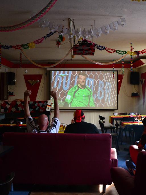 Eine überschaubare Gruppe von Fussballfans  schaut gemeinsam das Viertelfinalspiel Deutschland Frankreich im Juli 2014 in einer geschmückten Berliner Kneipe namens Brunnenquelle. Auf der Projektorleinwand ist Manuel Neuer eingeblendet.