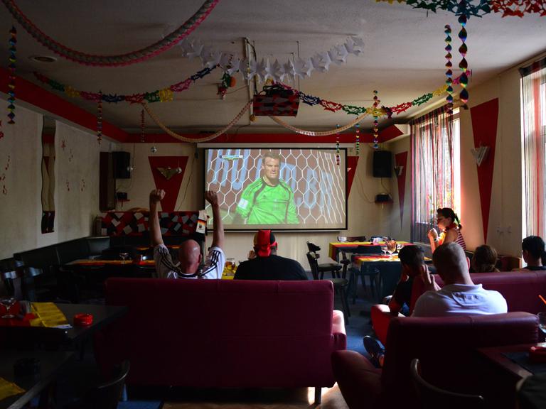 Eine überschaubare Gruppe von Fussballfans  schaut gemeinsam das Viertelfinalspiel Deutschland Frankreich im Juli 2014 in einer geschmückten Berliner Kneipe namens Brunnenquelle. Auf der Projektorleinwand ist Manuel Neuer eingeblendet.