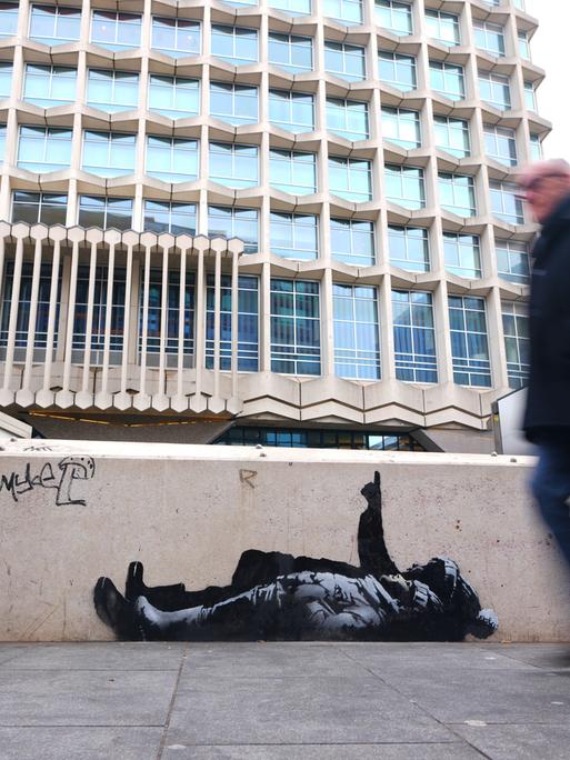 Auf einer niedrigen Mauer ist ein Graffiti von zwei Menschen abgebildet, die augenscheinlich auf dem Boden liegen und in den Himmel schauen. An der Mauer laufen mehrere Passanten vorbei