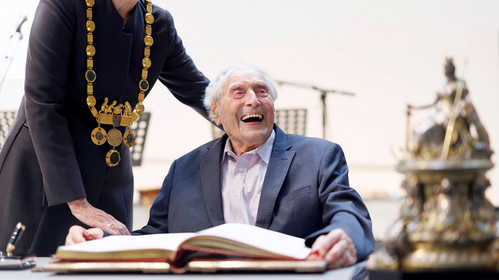 Ein sehr alter Mann sitzt lachend an einem großen Tisch. 