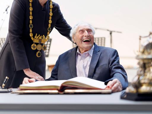 Ein sehr alter Mann sitzt lachend an einem großen Tisch. 